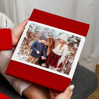 Hands holding a closed photo book with a red linen cover and photo dust jacket featuring an image of a mother with two children.