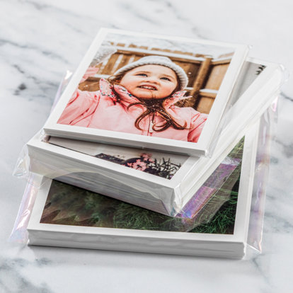 Three individual, cellophane wrapped stacks of signature photo prints. Each is a different size print. The photo seen on top is of a little girl in a hat and coat out in the snow.