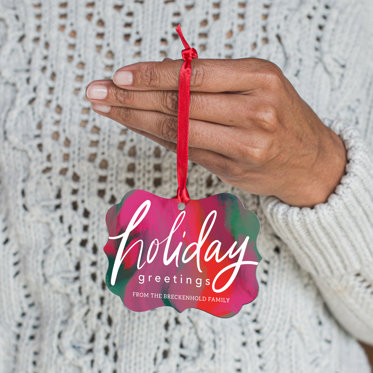 A hand holding the red ribbon of a holiday ornament featuring a colorful design and personalized text.