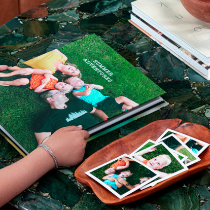 Hands holding a closed slip in photo album featuring a full image wrap cover of a mom and dad with their two kids lying on the grass with their heads together. A few photo prints of the same family are in a wooden tray next to the book.