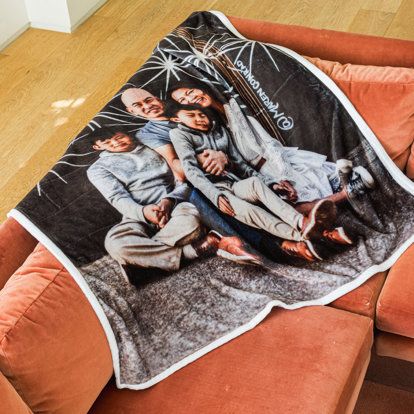 A photo blanket draped over the side of a couch. The blanket features one large photo of a family sitting outside in front of a black brick wall with fireworks painted on it.