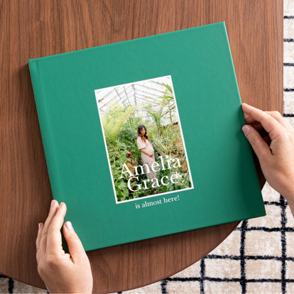 A closed photo book with teal cover and a single 4x6 photo of an expecting mother standing among plants in a greenhouse. The title reads 'Amelia Grace is almost here!'