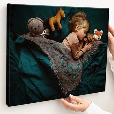 A canvas print wrap being mounted on a wall featuring a toddler asleep with his stuffed animals under a rich dark green blanket. 