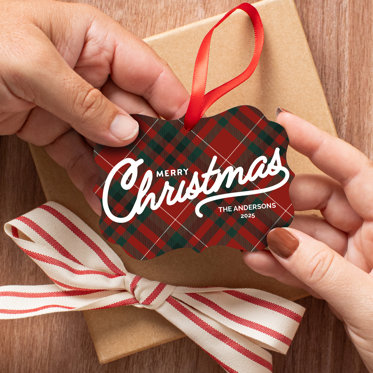 Hands exchanging a metal photo ornament that features a red and green plaid background and 'Merry Christmas' from 'The Andersons, 2025' in white text.