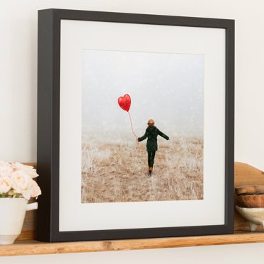 A framed photo print from Mpix in a black box frame with white matting and a photo of someone holding a heart shaped balloon for Valentines Day.