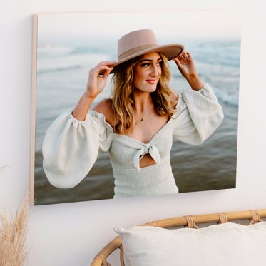 A wood photo print from Mpix mounted to a wall featuring a girl in a white dress and soft pink sun hat standing in front of a body of water for her senior photos.