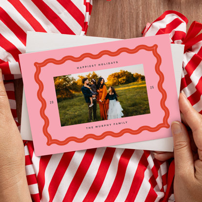 Hands holding a holiday card with a pink border surrounding a photo of a family in a field. The card reads 'Happiest Holidays 2025, The Murphy Family'.