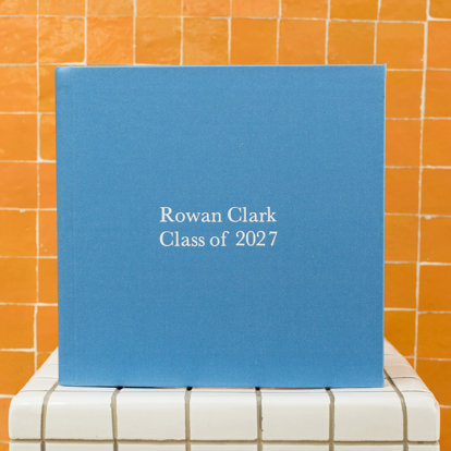 A closed custom fabric photo book with a bright blue cover and the title ‘Rowan Clark Class of 2027’ in silver debossing.