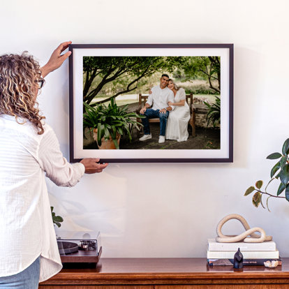 A woman hanging a framed photo print of a couple sitting on a wooden bench together with plants and trees around them. The frame is a flat black metal and includes a white window mat.
