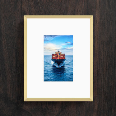 A framed print with a think gold frame and white window mat featuring a photo of a cargo ship in the ocean.