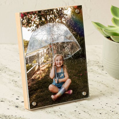 A vertical birch photo block displayed on a table and featuring a photo of a young girl sitting under an umbrella in the rain.