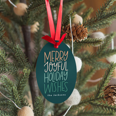 A metal holiday ornament hung on a Christmas tree with text that reads "Merry Joyful Holiday Wishes."