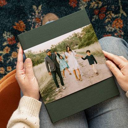 Hands holding a hardcover photo book with a green linen cover and a skinny dustjacket featuring a photo of a bride and groom wearing red, heart-shaped sunglasses. 