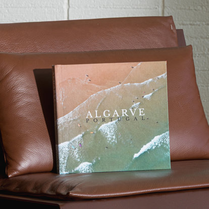 A closed custom fabric photo book featuring a full wraparound drone shot of people in the surf along on a sandy beach. The title of the book reads 'Algarve Portugal'.
