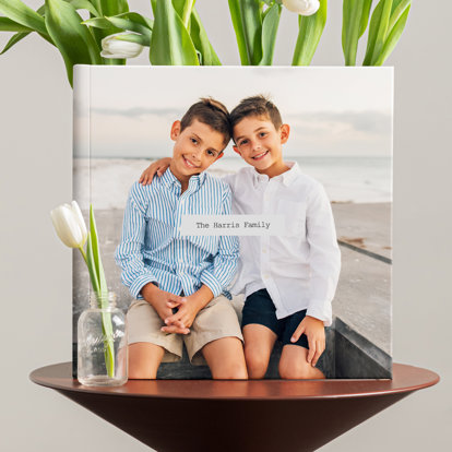 A closed custom cover photo book featuring a photo of two young brothers sitting on the beach. The book is displayed on a table next to a vase full of tulips.