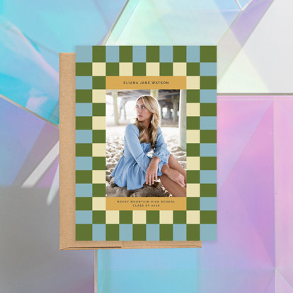 A graduation announcement featuring a 2026 graduating senior sitting on a beach under a pier with a checkered border.