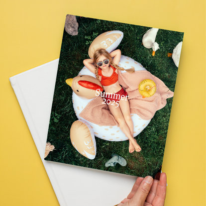 A closed hardcover photo book with a custom full-image cover featuring a little girl in a swimsuit and sunglasses lounging on an inflatable duck-shaped tube in the grass.