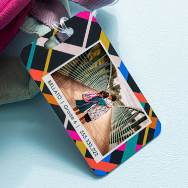 A metal luggage tag with a photo and personalized text on a colorful design that is attached to the handle of a backpack.