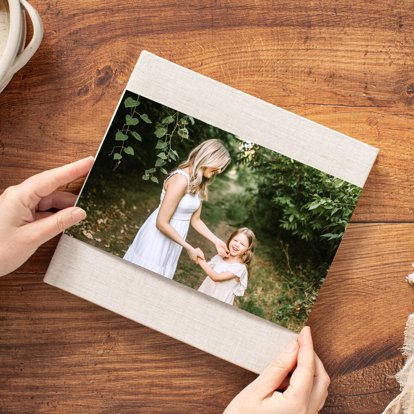 Hands holding a closed hardcover photo book with a tan linen cover and skinny dust jacket with a photo of a mother and daughter holding hands.