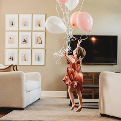 A toddler holding balloons in a living room with a grid of framed prints on the wall