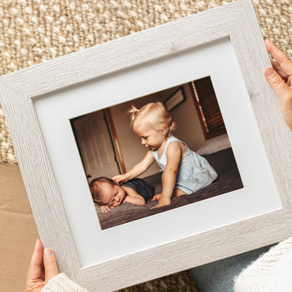a framed print with a white mat and a light wood frame frame featuring a young girl and her newest sibling