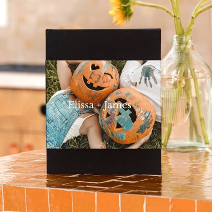 A photo book stood up on a counter next to flowers in a vase. The book has a black linen cover and skinny dust jacket featuring a Halloween-themed image and customized text.