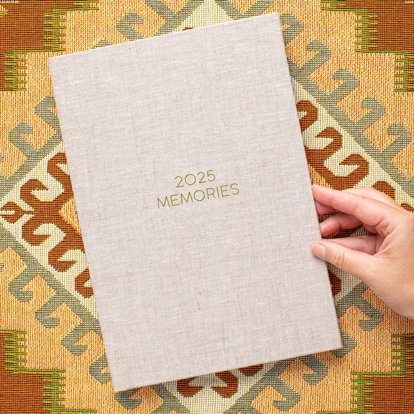 A closed 8x12 photo book with a sand color linen cover lying on a colorful cloth. The front cover has the text '2025 Memories.'