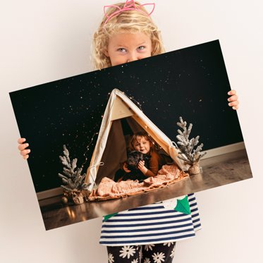Young girl holding a personalized metal print from Mpix of a toddler in a homemade tent next to two small holiday trees. 