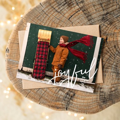 A Christmas photo card on top of a tan envelope laying on a natural wood end table.