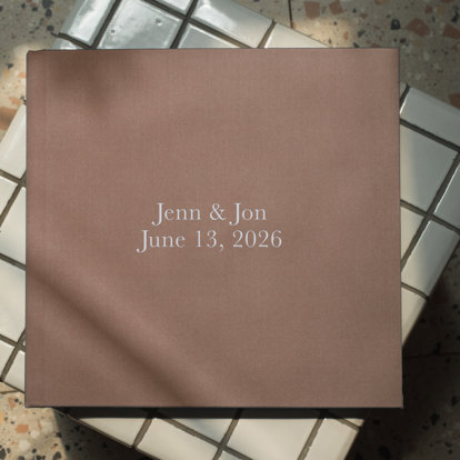 A closed custom fabric photo book with a brown fabric cover and the title ‘Jenn & Jon, June 13, 2026’ in silver debossing.
