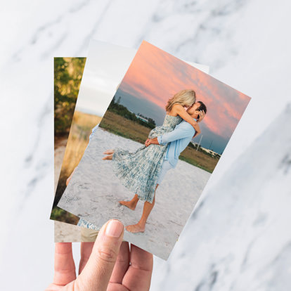 Hands holding a stack of 4x6 photo prints. The photo print on top features a husband and wife kissing at sunset on the beach. 