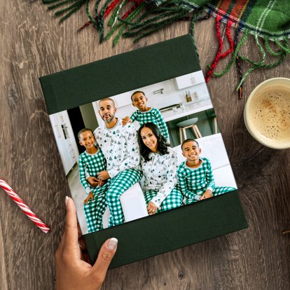 A hand holding the bottom left corner of a closed photo book with a dark green linen cover and photo dust jacket featuring an image of family in Christmas pajamas.