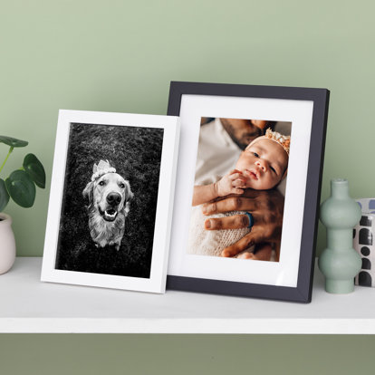 Two tabletop framed prints displayed on a table. One has a flat white frame and features a 4x6 black and white photo of a dog. The other is a flat black frame with white window mat and a 4x6 photo of a newborn baby.