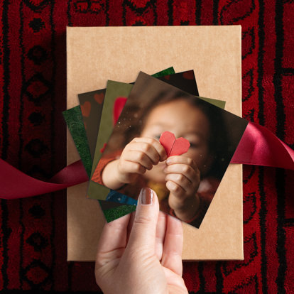 A hand holding a stack of photo prints. The top print shows a little girl holding a red origami heart.