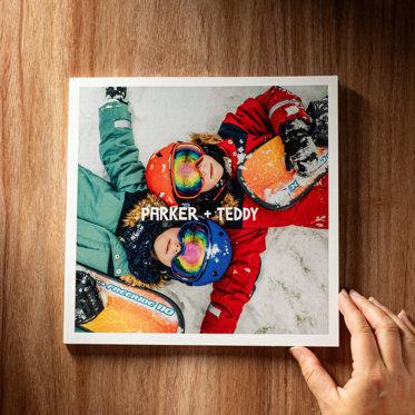 A hand holding a closed softcover photo book from Mpix on a wooden table featuring a photo of two children on the cover in showboarding gear laying in the snow with white text that reads "Parker + Teddy"