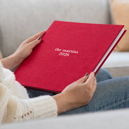 A woman sitting holding a closed hardcover photo book with a red linen cover and the text 'the martins 2026' in silver debossing.