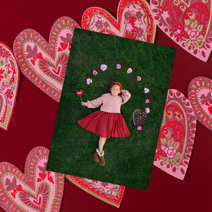 A 4x6 photo print of little girl lying in the grass with pink flowers circling her head. The photo is sitting on top of a string of valentine's heart cutouts.