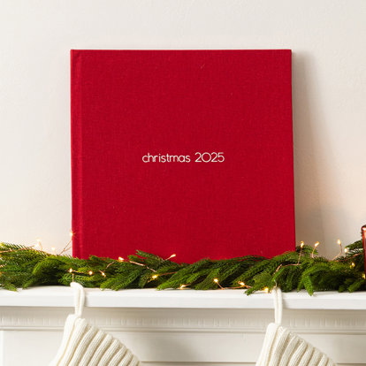 A closed photo book with a red linen cover and the text 'Christmas 2025' on the front. It's set on top of a mantel adorned with holiday garland.