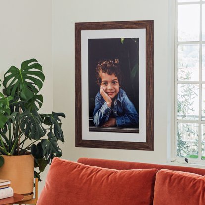 A framed photo print displayed on a home wall above a couch and next to a window and decorative plant. The print features a posed photo of a child. The frame is brown and has a white photo mat around the print.