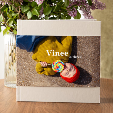 A closed linen hardcover photo book with a sand color linen cover and a skinny distjacket featuring a photo of a toddler lying on the sidewalk eating a giant lollipop. The title reads 'Vinee is three'. 