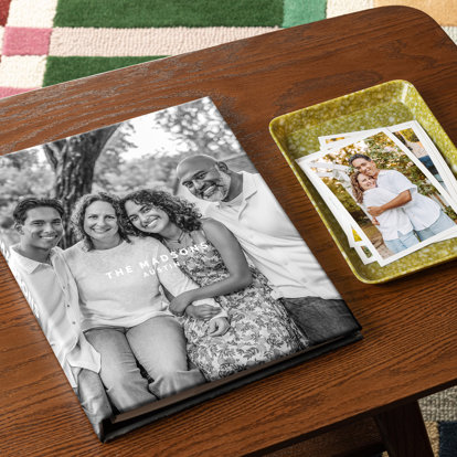 A closed photo book with a full wraparound cover featuring a photo of a couple and their adult children sitting on a bench in front of a tree. Next to the book is a tray containing various 4x6 prints of the same family.