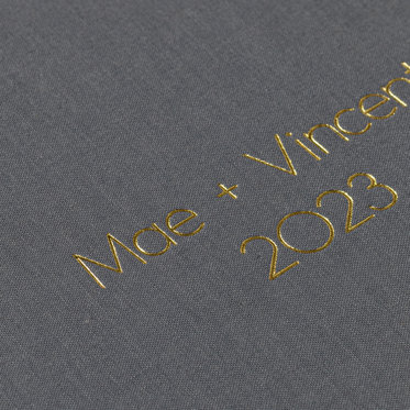 A close-up view of a wedding photo book with a graphite linen cover and gold foil debossing that reads "Mae + Vincent 2023."