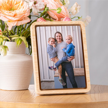 A framed metal print displayed on a table featuring a mom holding her two young boys, one in each arm.