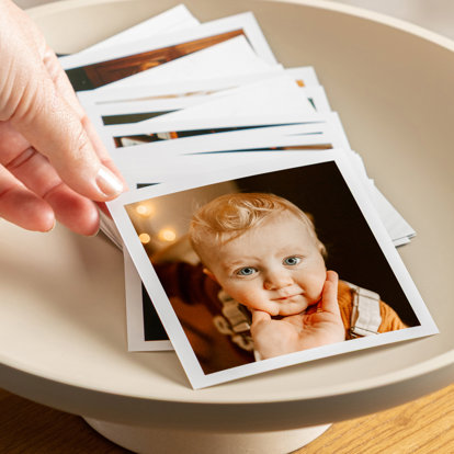 A decorative bowl full of 4x4" photo prints with a white border. The print on top features a closeup shot of a baby’s face.