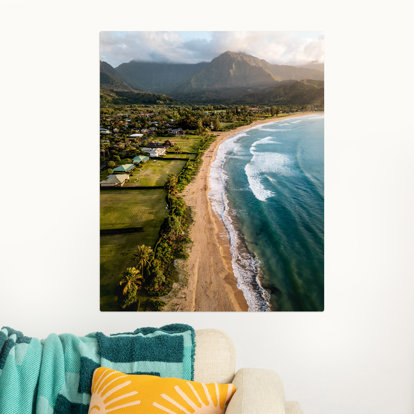 Large metal photo print hanging on a wall featuring a drone shot of a sandy beach, lush, green land, and mountains in the distance. 