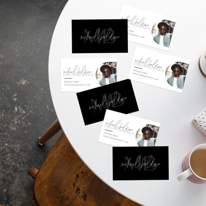 Business cards spread out on a table featuring a black front, and a white back with personalized details and a photoraph.