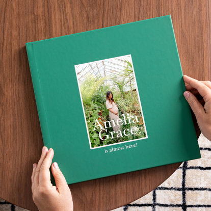 A closed photo book with teal cover and a single 4x6 photo of an expecting mother standing among plants in a greenhouse. The title reads 'Amelia Grace is almost here!'