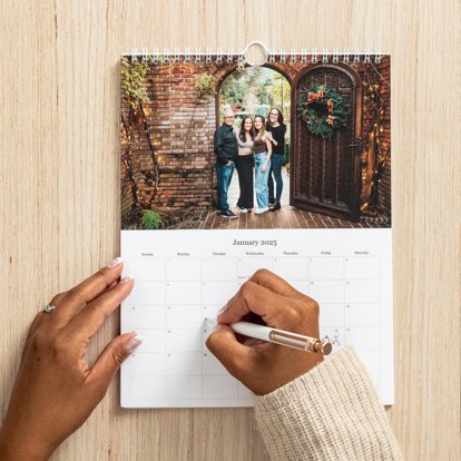 Hands using a pen to fill out the day grid of a wall calendar that also features a family photo on the top half.