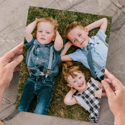 Hands holding an 8x10 photo print of three young siblings lying on the grass with their hands behind their heads.