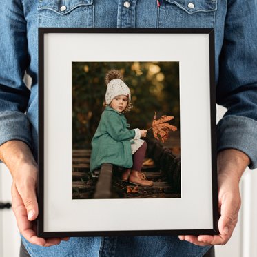 A framed print from Mpix with a 2" white mat and thin black metal frame being held, the image is of a toddler in a green rain coat sitting on railroad tracks holding a leaf from their fall family portraits.